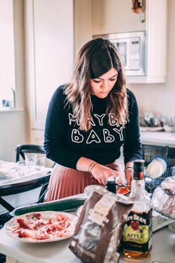 A woman with long brown hair is in the kitchen, focused on cutting a piece of fruit on a countertop. She wears a black sweater with the words 'Maybe Baby' printed on it. Surrounding her are various kitchen items, including a bottle of Jim Beam Apple on the table, a plate with slices of ham, a cucumber, and other food packaging.