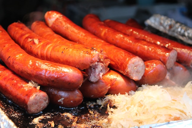 A pile of grilled sausages placed closely together. The sausages are glistening, indicating they are freshly cooked and juicy. There is a small portion of sauerkraut next to them, adding to the appetizing visual. Steam is lightly visible, suggesting heat and freshness.