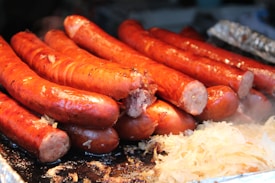 A pile of grilled sausages placed closely together. The sausages are glistening, indicating they are freshly cooked and juicy. There is a small portion of sauerkraut next to them, adding to the appetizing visual. Steam is lightly visible, suggesting heat and freshness.