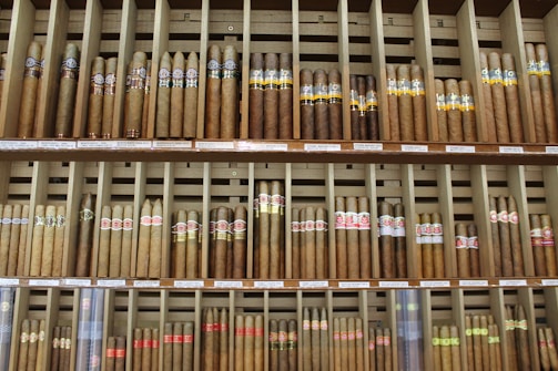 A neatly organized display of cigars in a wooden rack, with various brands and labels visible on the cigars. Each compartment contains an array of cigars, showing diverse shades of brown and different sizes. The setting suggests a retail environment for cigar enthusiasts.