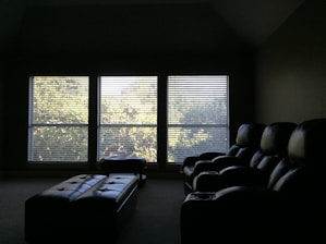 A dimly lit room features three large windows with blinds, allowing soft sunlight to filter through and cast a subtle glow on the room. In the foreground, there are plush, dark-colored recliner chairs arranged in a row, suggesting a home theater setup. A long, padded bench or ottoman sits in front of the chairs.