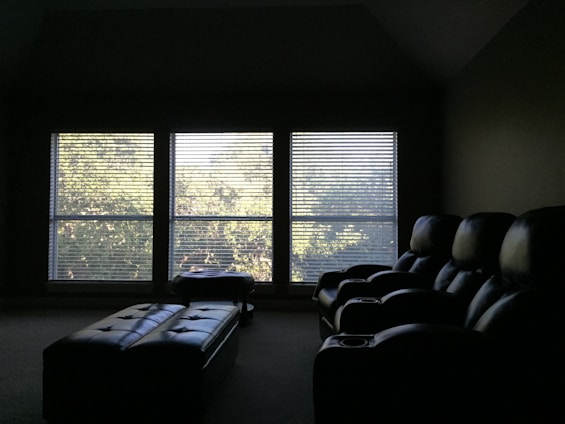 A dimly lit room features three large windows with blinds, allowing soft sunlight to filter through and cast a subtle glow on the room. In the foreground, there are plush, dark-colored recliner chairs arranged in a row, suggesting a home theater setup. A long, padded bench or ottoman sits in front of the chairs.
