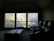 A dimly lit room features three large windows with blinds, allowing soft sunlight to filter through and cast a subtle glow on the room. In the foreground, there are plush, dark-colored recliner chairs arranged in a row, suggesting a home theater setup. A long, padded bench or ottoman sits in front of the chairs.
