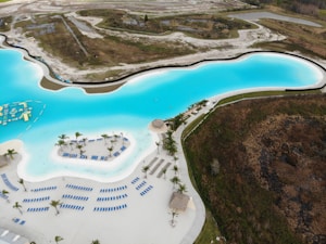 A large, clear blue lagoon surrounded by a landscaped area. The lagoon features a white sandy area dotted with rows of blue loungers and palm trees. Adjacent to the lagoon is a lush green expanse and a section of barren land. There are a few huts with thatched roofs along the beach.