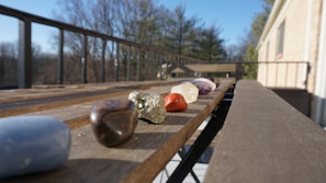 Several colorful stones and crystals are placed on a wooden surface, with a background of a balcony and trees. The stones are arranged in a line, with each stone varying in shape, size, and color.