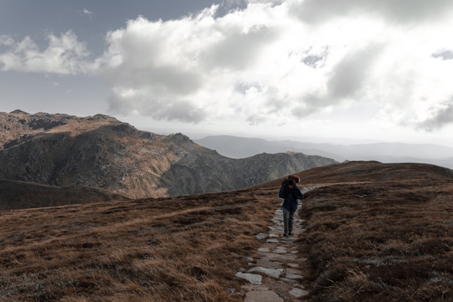 A lone hiker walks along a stone path on a grassy mountain ridge, with expansive views of rugged peaks under a sky filled with thick, scattered clouds.