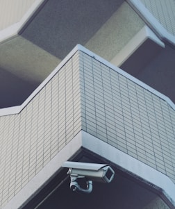 A security camera is mounted on the corner of a building with a tiled facade. The structure features geometric lines and shadows that create a modern and urban architectural feel.