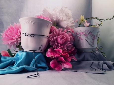 Two pots are creatively designed with wire figurines, surrounded by an arrangement of vibrant pink and white flowers. The scene features a soft textile background with blue and gray fabric draped beneath the pots, enhancing the floral display.