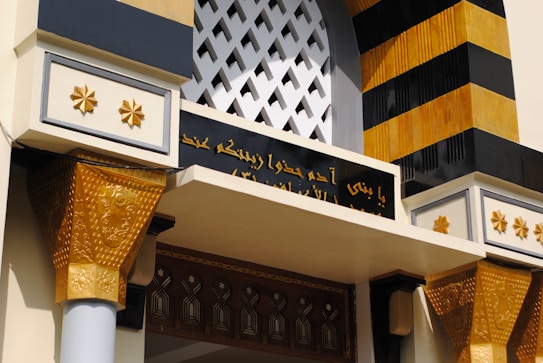 The image features an architectural detail with ornate patterns, including decorative columns with golden accents and geometric designs. There is an archway with latticework at the top, and a section of Arabic script is visible in black with gold lettering.