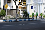 A group of women dressed in green and purple uniforms, some wearing hijabs, walks along a sidewalk beside a building with ornate geometric patterns and circular windows. The scene includes street signs indicating pedestrian crossings and well-maintained plants lining the path. The building's facade features decorative elements in white, black, and yellow.