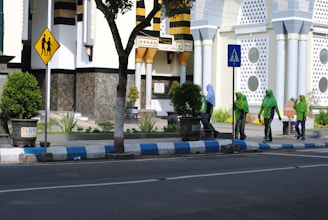 A group of women dressed in green and purple uniforms, some wearing hijabs, walks along a sidewalk beside a building with ornate geometric patterns and circular windows. The scene includes street signs indicating pedestrian crossings and well-maintained plants lining the path. The building's facade features decorative elements in white, black, and yellow.
