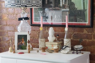 A decorative setup on a white dresser against a brick wall features a variety of items. There's a crystal lamp with a black and white geometric patterned lampshade, two tall pink candles in lace-covered holders, a bust sculpture, and several framed artworks. The surface is adorned with small ornamental pieces such as a bell, beads, and a small decorative box. The arrangement gives an eclectic and artistic vibe.
