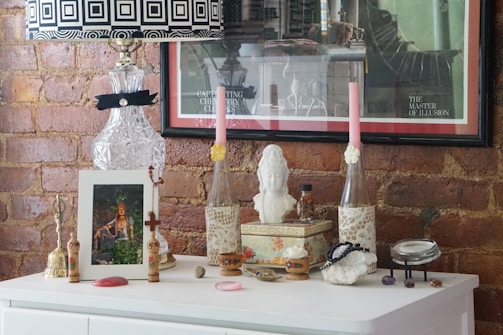 A decorative setup on a white dresser against a brick wall features a variety of items. There's a crystal lamp with a black and white geometric patterned lampshade, two tall pink candles in lace-covered holders, a bust sculpture, and several framed artworks. The surface is adorned with small ornamental pieces such as a bell, beads, and a small decorative box. The arrangement gives an eclectic and artistic vibe.