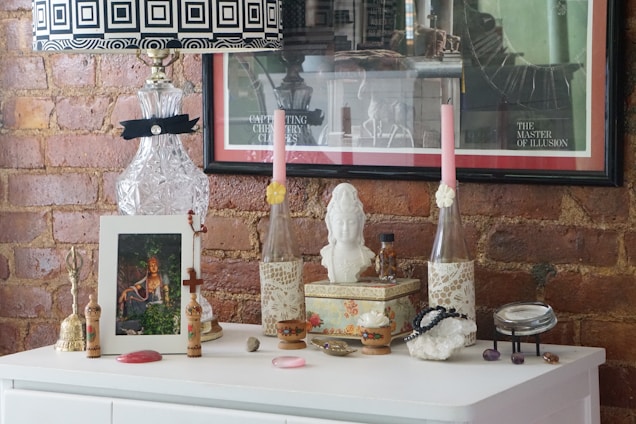 A decorative setup on a white dresser against a brick wall features a variety of items. There's a crystal lamp with a black and white geometric patterned lampshade, two tall pink candles in lace-covered holders, a bust sculpture, and several framed artworks. The surface is adorned with small ornamental pieces such as a bell, beads, and a small decorative box. The arrangement gives an eclectic and artistic vibe.