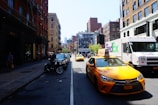 A busy urban street scene with yellow taxis and a FedEx delivery truck. The street is lined with brick buildings on both sides, some with fire escapes. Cars and motorcycles are parked along the sidewalk, and a large mural can be seen in the background.