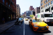 A busy urban street scene with yellow taxis and a FedEx delivery truck. The street is lined with brick buildings on both sides, some with fire escapes. Cars and motorcycles are parked along the sidewalk, and a large mural can be seen in the background.