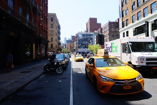 A busy urban street scene with yellow taxis and a FedEx delivery truck. The street is lined with brick buildings on both sides, some with fire escapes. Cars and motorcycles are parked along the sidewalk, and a large mural can be seen in the background.