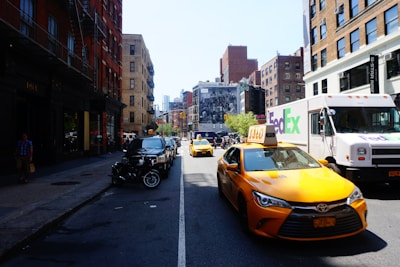 A busy urban street scene with yellow taxis and a FedEx delivery truck. The street is lined with brick buildings on both sides, some with fire escapes. Cars and motorcycles are parked along the sidewalk, and a large mural can be seen in the background.