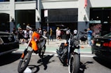 Two motorcycles are parked on a street in front of a group of people standing near a building entrance. The building has tall windows, and there are two cars on either side of the motorcycles. The people appear to be socializing or waiting, with some looking at their phones.