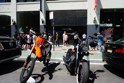 Two motorcycles are parked on a street in front of a group of people standing near a building entrance. The building has tall windows, and there are two cars on either side of the motorcycles. The people appear to be socializing or waiting, with some looking at their phones.