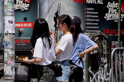 Three people stand near a utility pole covered with graffiti and posters. Behind them is a wall with a large advertisement for a safety fire sprinkler company. Two women wear casual clothes and carry shopping bags, and one wears a baseball cap. The scene is urban with elements of street art and advertising.