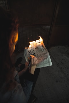 A person is holding a newspaper that is partially on fire. The setting appears to be dimly lit, possibly indoors, with a focus on the flames and the newspaper. The newspaper shows various comic strips and is being held with one hand while a pen is held in the other hand.