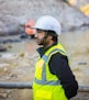 A person wearing a white hard hat, safety goggles, and a bright yellow high-visibility vest is standing at a construction site. The background shows blurred construction equipment and rocky terrain, indicating an outdoor work environment.