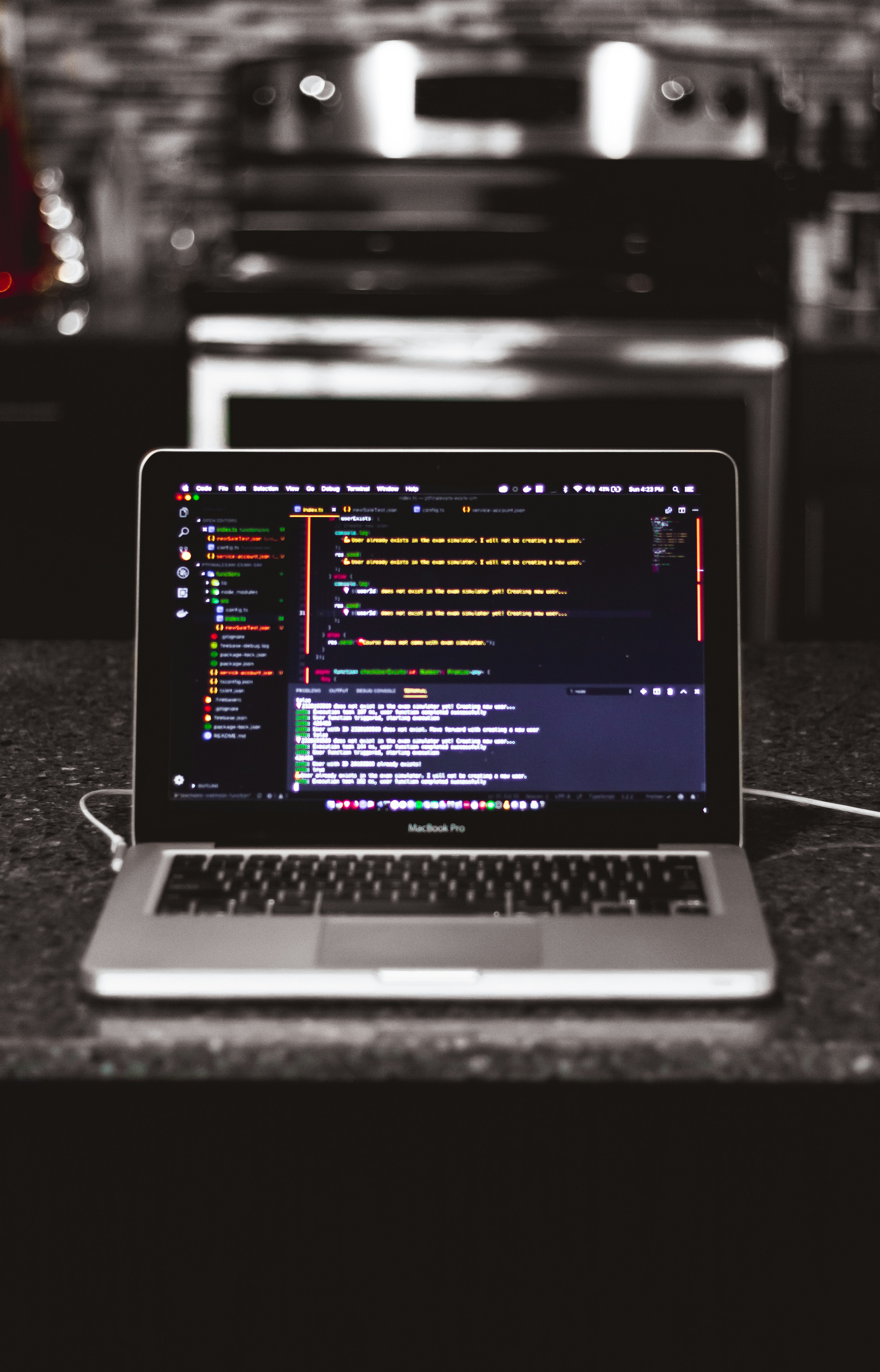 A laptop is placed on a dark granite surface. The screen displays coding software with lines of colorful code. The background features a modern kitchen with a blurred stovetop and backsplash.