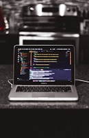 A laptop is placed on a dark granite surface. The screen displays coding software with lines of colorful code. The background features a modern kitchen with a blurred stovetop and backsplash.