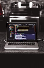 A laptop is placed on a dark granite surface. The screen displays coding software with lines of colorful code. The background features a modern kitchen with a blurred stovetop and backsplash.