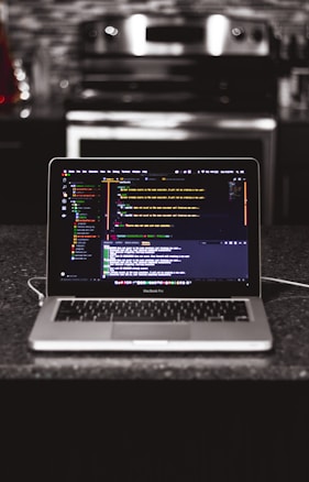 A laptop is placed on a dark granite surface. The screen displays coding software with lines of colorful code. The background features a modern kitchen with a blurred stovetop and backsplash.
