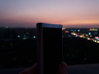 Close-up of a hand holding a compact Bluetooth device against a blurred cityscape.