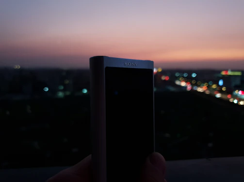 Hands holding a compact Village Sounds speaker against a backdrop of city lights at dusk.