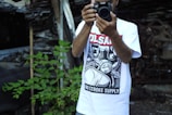 A person is holding a camera, wearing a graphic t-shirt with bold black and white comic-style artwork and text in red and black. Green plants are visible to the side, and there is a backdrop of a cluttered or possibly industrial or scrap setting.