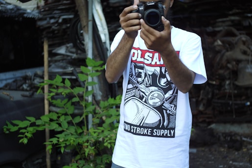 A person is holding a camera, wearing a graphic t-shirt with bold black and white comic-style artwork and text in red and black. Green plants are visible to the side, and there is a backdrop of a cluttered or possibly industrial or scrap setting.
