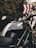 A sleek black Ducati motorcycle prominently displayed, with a silver stripe running down the middle of the fuel tank. The background includes some green foliage and a road sign with red and white diagonal stripes. The motorcycle's side mirror and part of the handlebars are visible.
