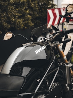 A sleek black Ducati motorcycle prominently displayed, with a silver stripe running down the middle of the fuel tank. The background includes some green foliage and a road sign with red and white diagonal stripes. The motorcycle's side mirror and part of the handlebars are visible.