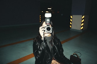 A person dressed in a dark leather jacket is holding a camera with a flash attached, aiming it directly at the viewer. The setting appears to be an indoor parking garage with visible striped pillars in the background, providing an industrial and moody atmosphere.