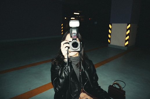 A person dressed in a dark leather jacket is holding a camera with a flash attached, aiming it directly at the viewer. The setting appears to be an indoor parking garage with visible striped pillars in the background, providing an industrial and moody atmosphere.