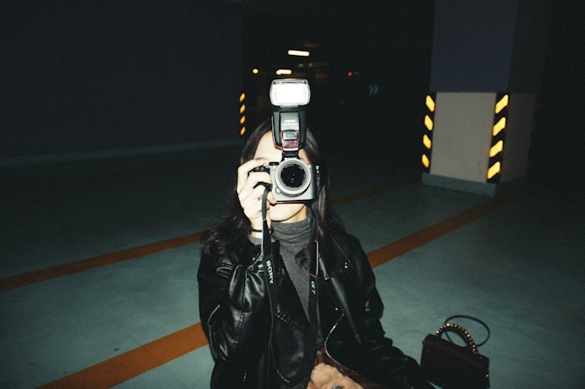 A person dressed in a dark leather jacket is holding a camera with a flash attached, aiming it directly at the viewer. The setting appears to be an indoor parking garage with visible striped pillars in the background, providing an industrial and moody atmosphere.