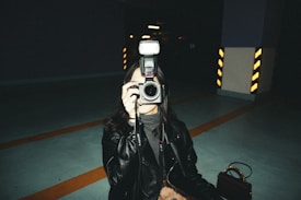 A person dressed in a dark leather jacket is holding a camera with a flash attached, aiming it directly at the viewer. The setting appears to be an indoor parking garage with visible striped pillars in the background, providing an industrial and moody atmosphere.