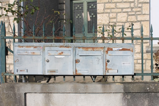A set of four old, rusted metal mailboxes is mounted on a stone wall. Behind the mailboxes, there is a green metal fence with spiked tops and some sparse, leafless branches woven through it. The background features a stone building with a window that has decorative metal grills.