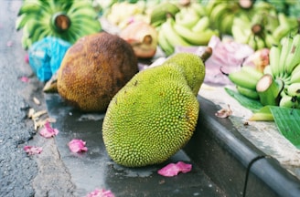 A variety of fresh fruits displayed on a market stall. In the foreground, two jackfruits, one ripe and brown, the other green, are positioned on a dark surface. Surrounding them are several bunches of bananas and other fruits scattered with pink petals on the ground.