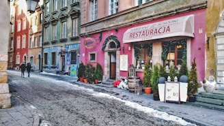 A charming street scene features colorful historic buildings with intricate architectural details. A cozy restaurant with a sign displaying 'Restauracja' is adorned with decorative plants and lanterns at the entrance. Two people walk along the cobblestone pathway, with a few patches of snow scattered along the sides.