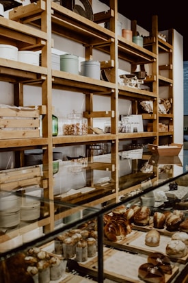 A bakery display with shelves filled with various containers and wooden boxes. The counter is lined with a variety of freshly baked goods, including muffins and pastries. The setting has a rustic and cozy atmosphere with wooden shelves and glass display cases.