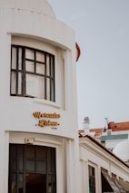 A white building with large black-framed windows and a sign reading 'Mercado Lisboa' in stylish yellow letters. The architecture has a modern yet classic feel, with a domed roof and columns. A clear sky forms the background, and part of a red-tiled roof is visible.