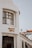 A white building with large black-framed windows and a sign reading 'Mercado Lisboa' in stylish yellow letters. The architecture has a modern yet classic feel, with a domed roof and columns. A clear sky forms the background, and part of a red-tiled roof is visible.