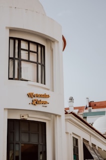A white building with large black-framed windows and a sign reading 'Mercado Lisboa' in stylish yellow letters. The architecture has a modern yet classic feel, with a domed roof and columns. A clear sky forms the background, and part of a red-tiled roof is visible.