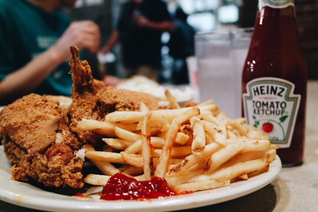 A close-up of a plate filled with crispy fried chicken and a large pile of golden french fries accompanied by a dollop of ketchup. In the background, there are blurred figures of people. On the table, there is a bottle of Heinz tomato ketchup and two glasses of water.
