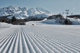 Groomed ski slopes stretch towards rugged, snow-capped mountains under a clear blue sky. Cable cars traverse overhead, positioned to the right, while trees sparsely line the lower slopes, contrasting against the pristine white snow.